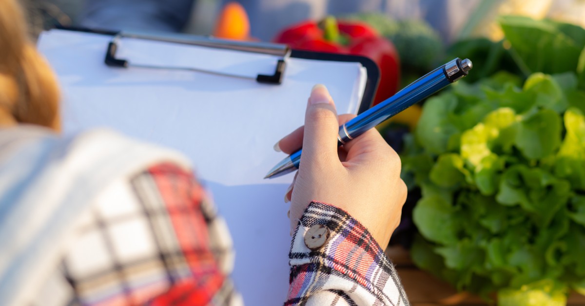 A close-up of a woman holding a clipboard and a pen. Behind the clipboard are multiple vegetables and other plants.