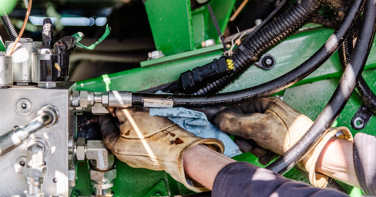 A mechanic wearing tan gloves working inside of a tractor. He cleans the hose with a blue washcloth.