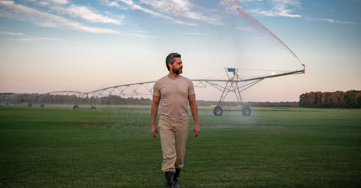 A worker wearing boots is walking towards the camera in a large field of green grass with a large irrigation system.