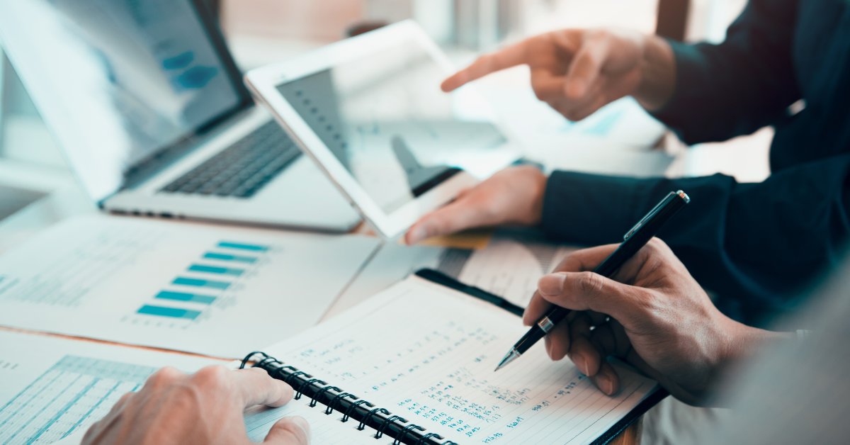 A person writes numbers in a small notebook while another person shows them a charts on a laptop and tablet.