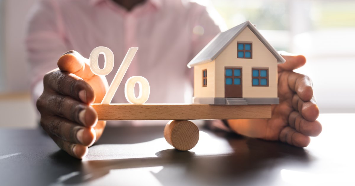 A person holds a small wooden seesaw. A percent symbol is on one end, and a small home is on the other.