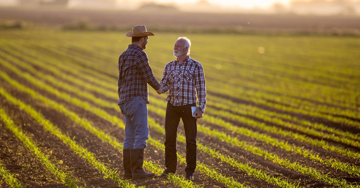 A large plot of farmland with a worker in boots and a hat shakes hands with another man holding a tablet.