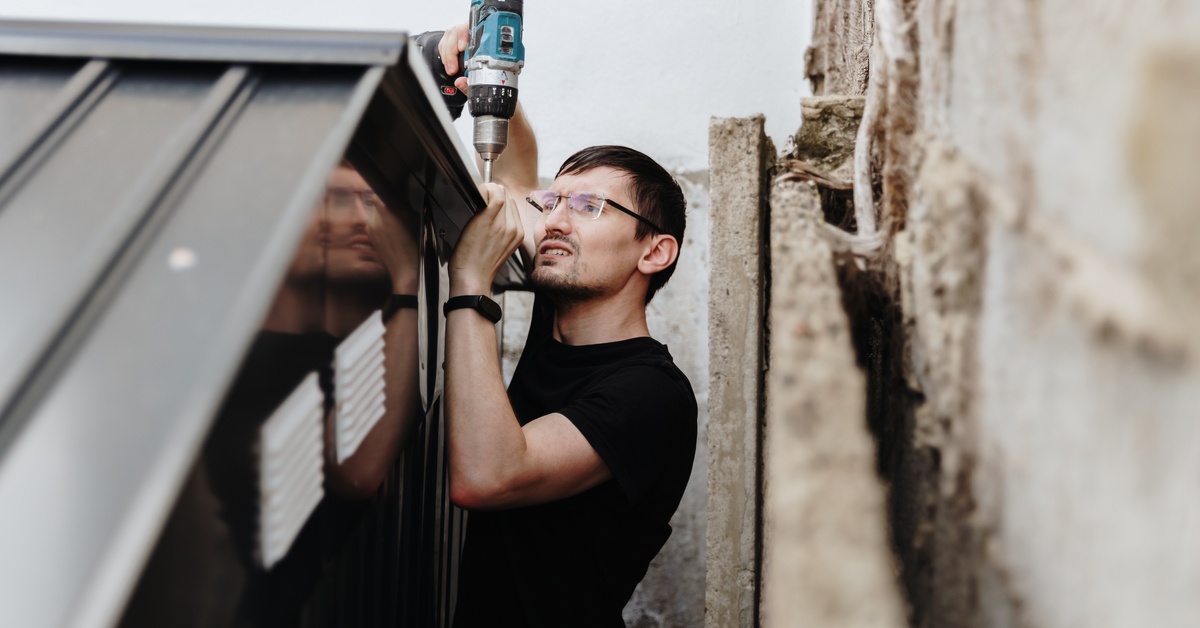 A man drilling into the top of a small metal farming structure. It is backed up close to a tall stone wall.