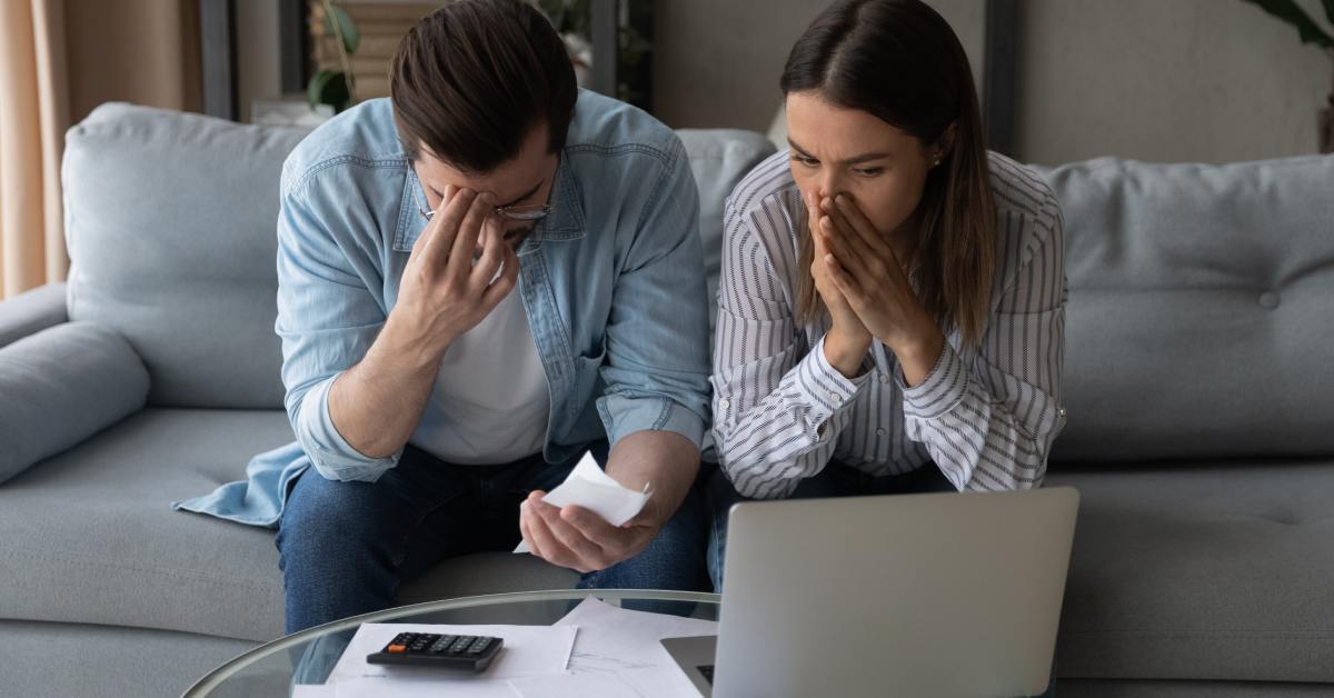 Two distressed people sitting on the couch, looking at some paperwork and a laptop. They also have a calculator nearby.