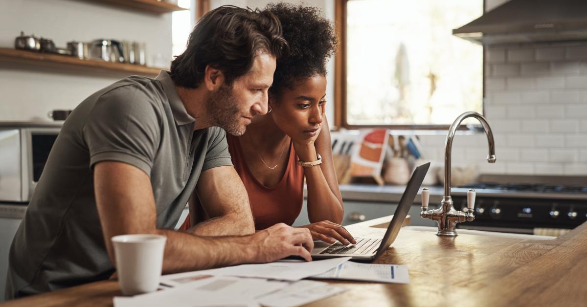Two people looking at a laptop together at their kitchen island. They have some paperwork spread out next to them.