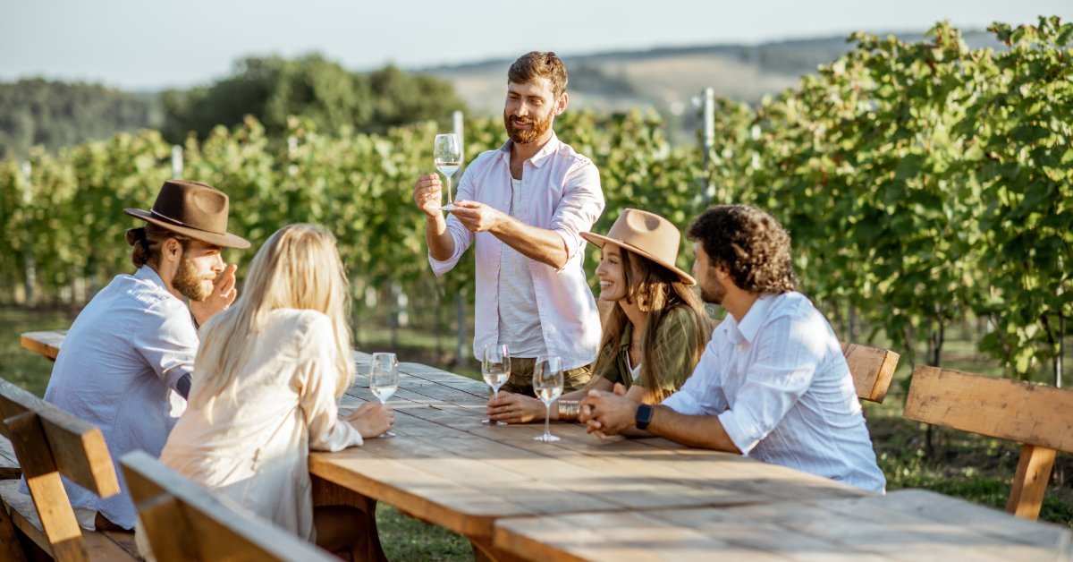 A person holds a wine glass in front of a table in an outdoor vineyard. Four people sit at the table with drinks.